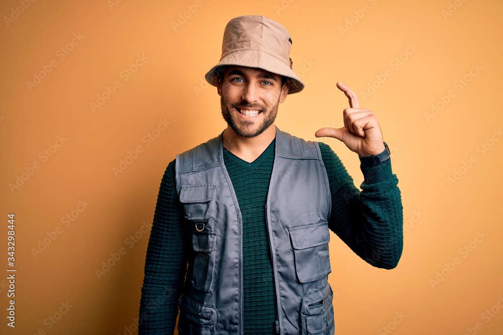 Handsome tourist man with beard on vacation wearing explorer hat over yellow background smiling and confident gesturing with hand doing small size sign with fingers looking and the camera. Measure Handsome tourist man with beard on vacation wearing explorer hat over yellow background smiling and confident gesturing with hand doing small size sign with fingers looking and the camera. Measure