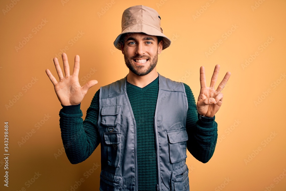 Handsome tourist man with beard on vacation wearing explorer hat over yellow background showing and pointing up with fingers number eight while smiling confident and happy.