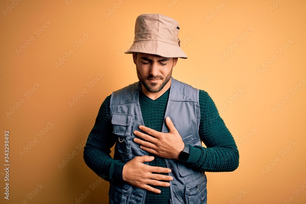 Handsome tourist man with beard on vacation wearing explorer hat over yellow background with hand on stomach because indigestion, painful illness feeling unwell. Ache concept. Handsome tourist man with beard on vacation wearing explorer hat over yellow background with hand on stomach because indigestion, painful illness feeling unwell. Ache concept.