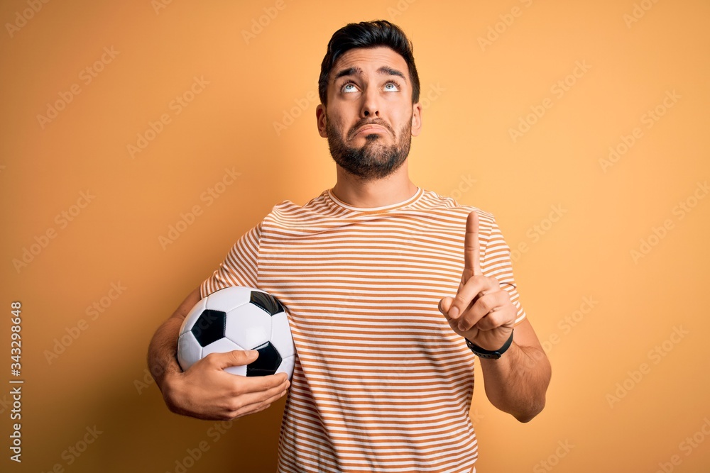 Handsome player man with beard playing soccer holding footballl ball over yellow background Pointing up looking sad and upset, indicating direction with fingers, unhappy and depressed. Handsome player man with beard playing soccer holding footballl ball over yellow background Pointing up looking sad and upset, indicating direction with fingers, unhappy and depressed.