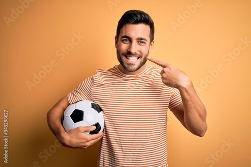 Fototapeta Naklejka Na Ścianę i Meble -  Handsome player man with beard playing soccer holding footballl ball over yellow background smiling cheerful showing and pointing with fingers teeth and mouth. Dental health concept.