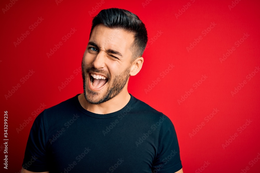 Young handsome man wearing casual black t-shirt standing over isolated red background winking looking at the camera with sexy expression, cheerful and happy face. Young handsome man wearing casual black t-shirt standing over isolated red background winking looking at the camera with sexy expression, cheerful and happy face.