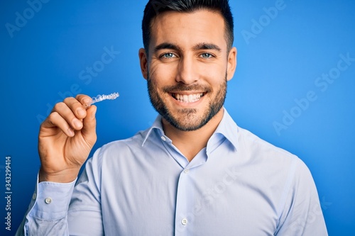 Young handsome man holding dental aligner tooth correction over blue background with a happy face standing and smiling with a confident smile showing teeth