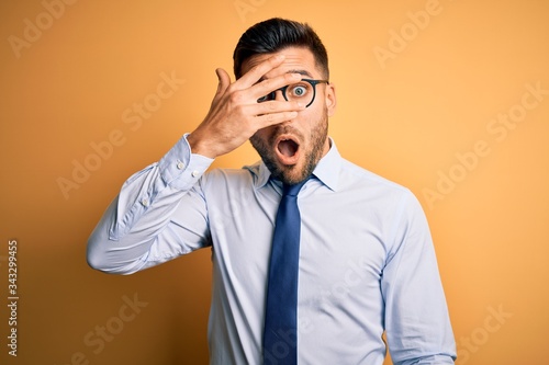 Young handsome businessman wearing tie and glasses standing over yellow background peeking in shock covering face and eyes with hand, looking through fingers with embarrassed expression. © Krakenimages.com