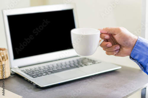 Business people hold a cup of coffee and use a laptop on a wooden table beside the window to work at home, the background is close up.