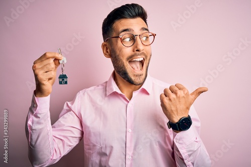 Young real estate business man holding new house keys over pink background pointing and showing with thumb up to the side with happy face smiling