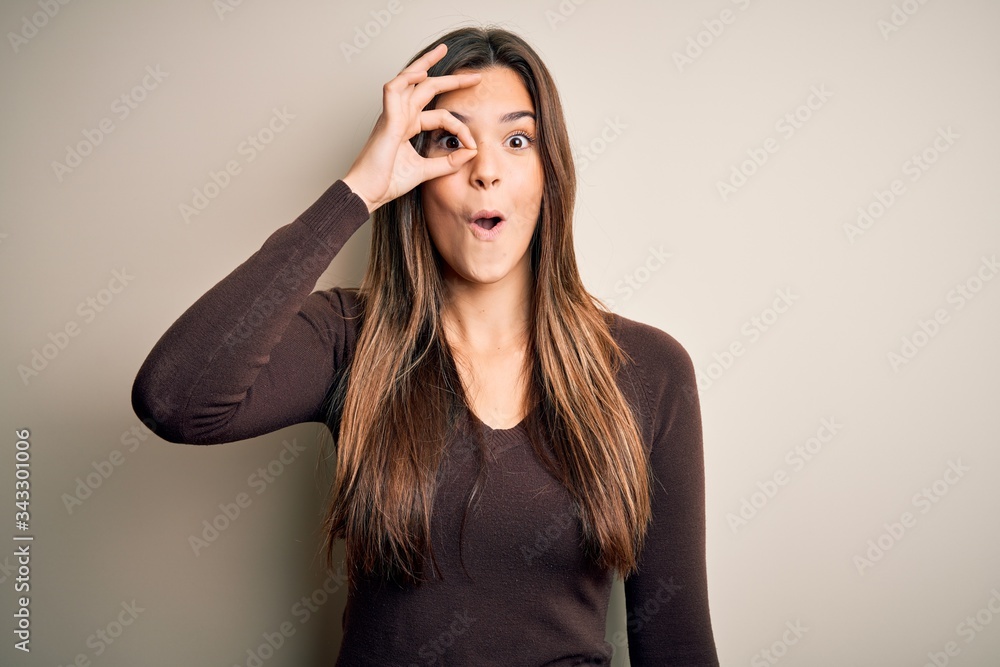 Young beautiful girl wearing casual sweater standing over isolated white background doing ok gesture shocked with surprised face, eye looking through fingers. Unbelieving expression. Young beautiful girl wearing casual sweater standing over isolated white background doing ok gesture shocked with surprised face, eye looking through fingers. Unbelieving expression.