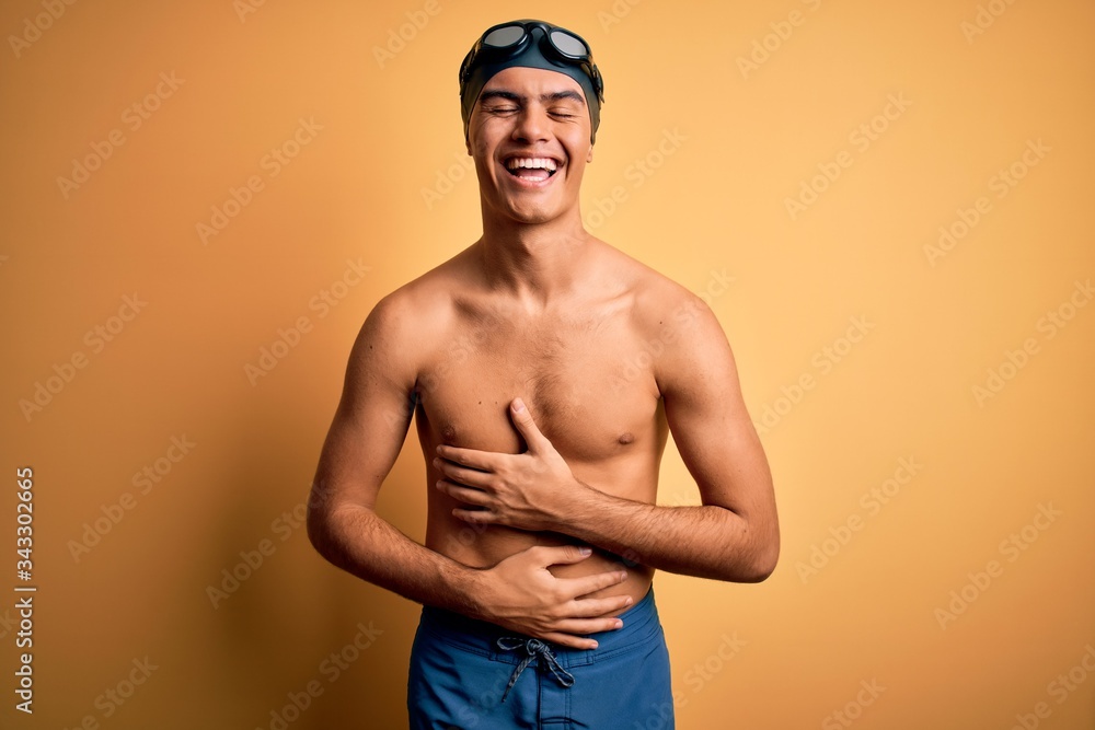 Young handsome man shirtless wearing swimsuit and swim cap over isolated yellow background smiling and laughing hard out loud because funny crazy joke with hands on body.