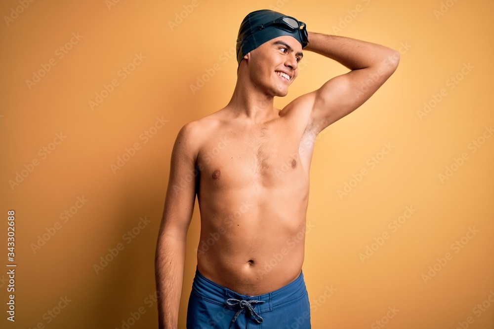 Young handsome man shirtless wearing swimsuit and swim cap over isolated yellow background smiling confident touching hair with hand up gesture, posing attractive and fashionable