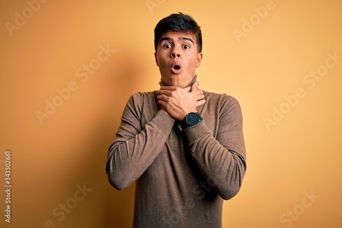 Young handsome man wearing casual turtleneck sweater over isolated yellow background shouting and suffocate because painful strangle. Health problem. Asphyxiate and suicide concept.