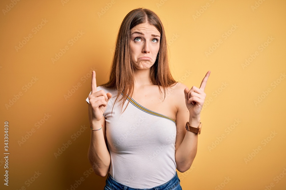 Young beautiful redhead woman wearing casual t-shirt over isolated yellow background Pointing up looking sad and upset, indicating direction with fingers, unhappy and depressed. Young beautiful redhead woman wearing casual t-shirt over isolated yellow background Pointing up looking sad and upset, indicating direction with fingers, unhappy and depressed.