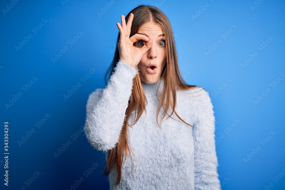 Young beautiful redhead woman wearing casual sweater over isolated blue background doing ok gesture shocked with surprised face, eye looking through fingers. Unbelieving expression. Young beautiful redhead woman wearing casual sweater over isolated blue background doing ok gesture shocked with surprised face, eye looking through fingers. Unbelieving expression.