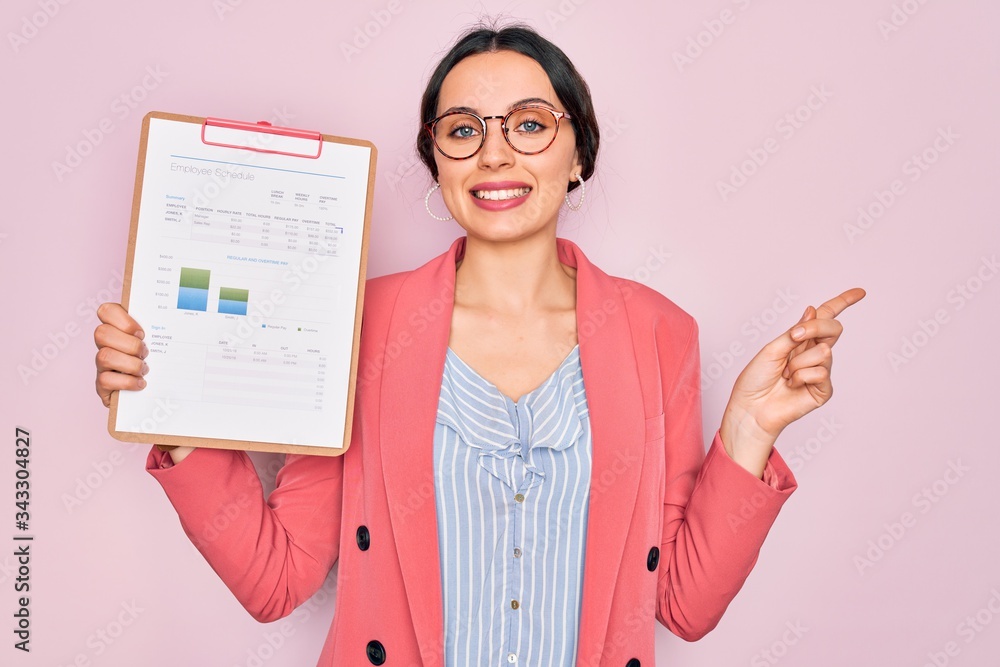 Young beautiful business woman with blue eyes wearing glasses holding clipboard very happy pointing with hand and finger to the side Young beautiful business woman with blue eyes wearing glasses holding clipboard very happy pointing with hand and finger to the side