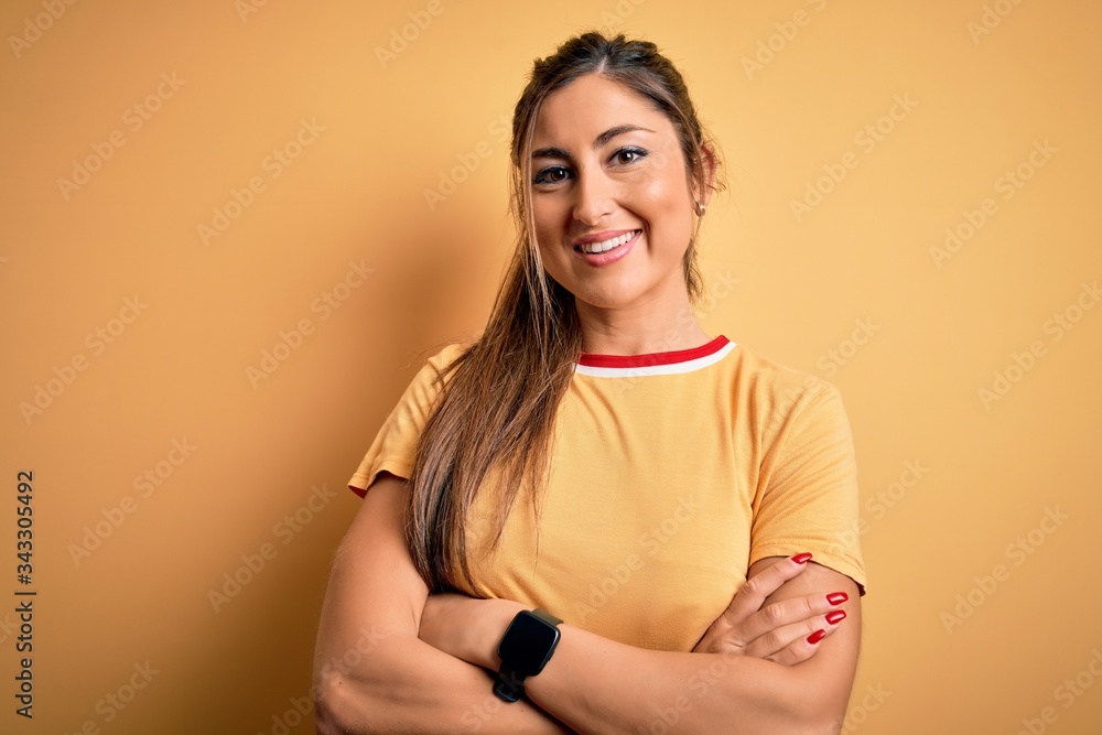 Obraz premium Young beautiful athletic woman wearing casual t-shirt and ponytail over yellow background happy face smiling with crossed arms looking at the camera. Positive person.