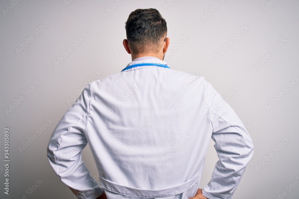 Young doctor man with blue eyes wearing medical coat and stethoscope over isolated background standing backwards looking away with arms on body Young doctor man with blue eyes wearing medical coat and stethoscope over isolated background standing backwards looking away with arms on body