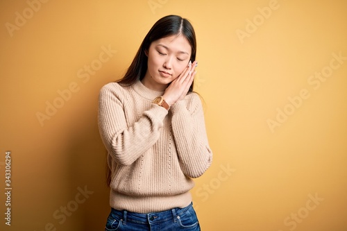 Young beautiful asian woman wearing casual sweater over yellow isolated background sleeping tired dreaming and posing with hands together while smiling with closed eyes.