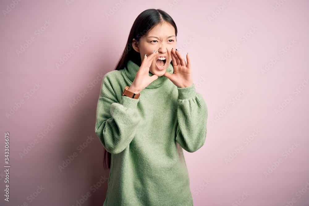 Young beautiful asian woman wearing green winter sweater over pink solated background Shouting angry out loud with hands over mouth Young beautiful asian woman wearing green winter sweater over pink solated background Shouting angry out loud with hands over mouth