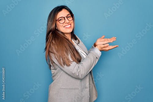 Young hispanic business woman wearing glasses standing over blue isolated background pointing aside with hands open palms showing copy space, presenting advertisement smiling excited happy photo