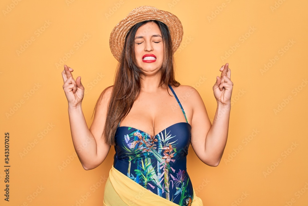 Young hispanic woman wearing summer hat and swimsuit over yellow background gesturing finger crossed smiling with hope and eyes closed. Luck and superstitious concept. Young hispanic woman wearing summer hat and swimsuit over yellow background gesturing finger crossed smiling with hope and eyes closed. Luck and superstitious concept.
