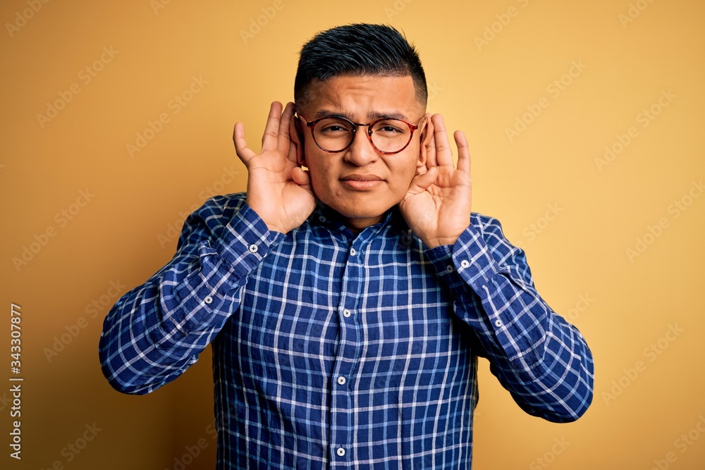 Young handsome latin man wearing casual shirt and glasses over yellow background Trying to hear both hands on ear gesture, curious for gossip. Hearing problem, deaf Young handsome latin man wearing casual shirt and glasses over yellow background Trying to hear both hands on ear gesture, curious for gossip. Hearing problem, deaf