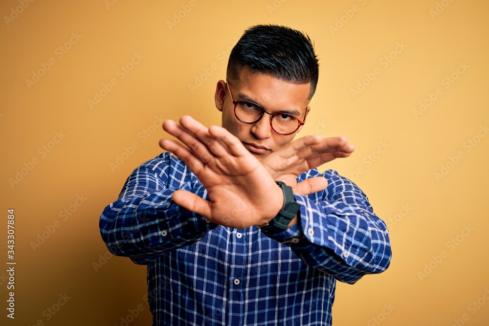 Young handsome latin man wearing casual shirt and glasses over yellow background Rejection expression crossing arms doing negative sign, angry face Young handsome latin man wearing casual shirt and glasses over yellow background Rejection expression crossing arms doing negative sign, angry face
