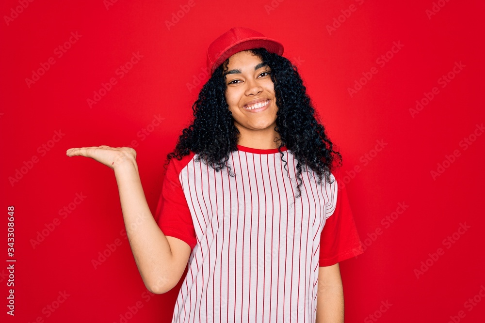 Young african american curly sportswoman wearing baseball cap and striped t-shirt smiling cheerful presenting and pointing with palm of hand looking at the camera. Young african american curly sportswoman wearing baseball cap and striped t-shirt smiling cheerful presenting and pointing with palm of hand looking at the camera.