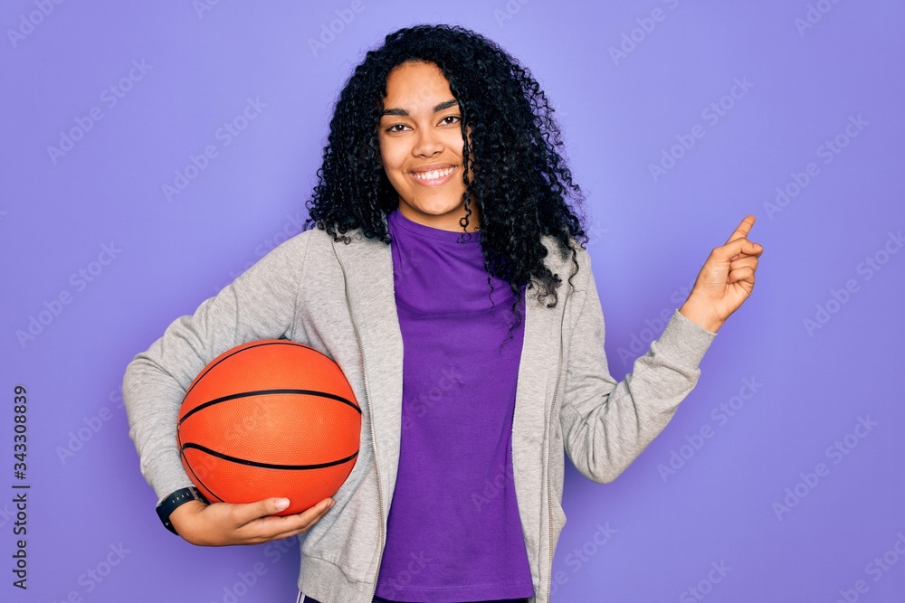African american curly sportswoman doing sport holding basketball ball over purple background very happy pointing with hand and finger to the side