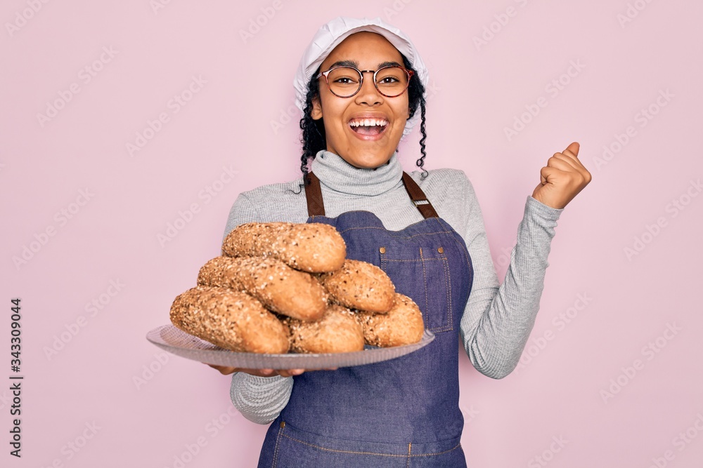 Young african american baker woman wearing apron holding wholemeal bread with cereal screaming proud and celebrating victory and success very excited, cheering emotion Young african american baker woman wearing apron holding wholemeal bread with cereal screaming proud and celebrating victory and success very excited, cheering emotion
