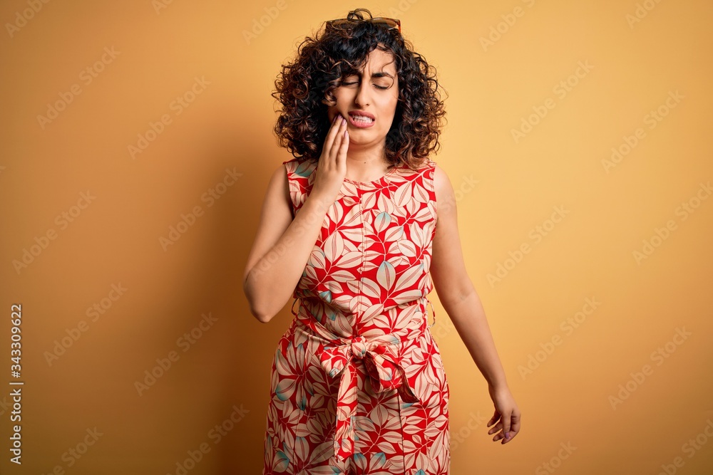Young beautiful curly arab woman on vacation wearing summer floral dress and sunglasses touching mouth with hand with painful expression because of toothache or dental illness on teeth. Dentist
