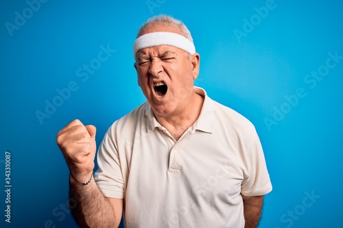 Middle age hoary sportsman doing sport wearing sportswear over isolated blue background angry and mad raising fist frustrated and furious while shouting with anger. Rage and aggressive concept.