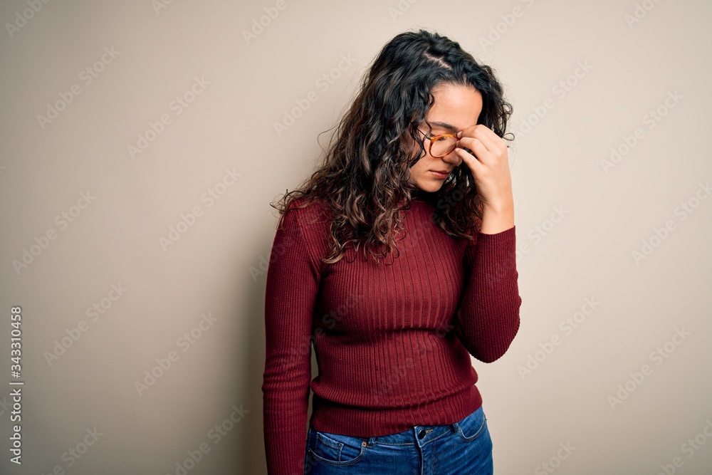 Beautiful woman with curly hair wearing casual sweater and glasses over white background tired rubbing nose and eyes feeling fatigue and headache. Stress and frustration concept. Beautiful woman with curly hair wearing casual sweater and glasses over white background tired rubbing nose and eyes feeling fatigue and headache. Stress and frustration concept.