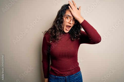 Beautiful woman with curly hair wearing casual sweater and glasses over white background surprised with hand on head for mistake, remember error. Forgot, bad memory concept.
