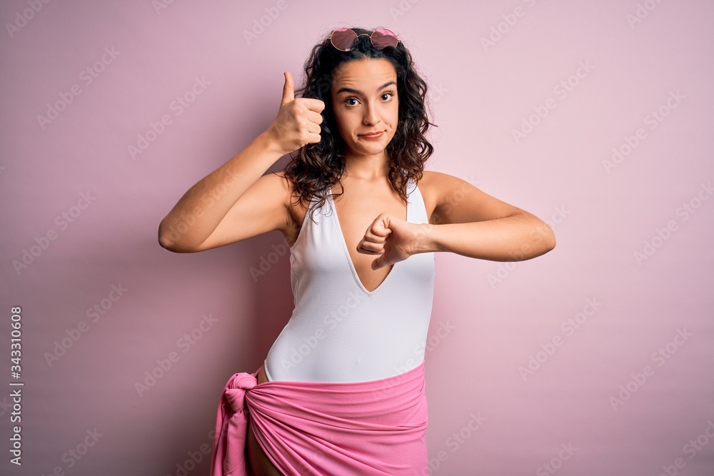 Beautiful woman with curly hair on vacation wearing white swimsuit over pink background Doing thumbs up and down, disagreement and agreement expression. Crazy conflict Beautiful woman with curly hair on vacation wearing white swimsuit over pink background Doing thumbs up and down, disagreement and agreement expression. Crazy conflict