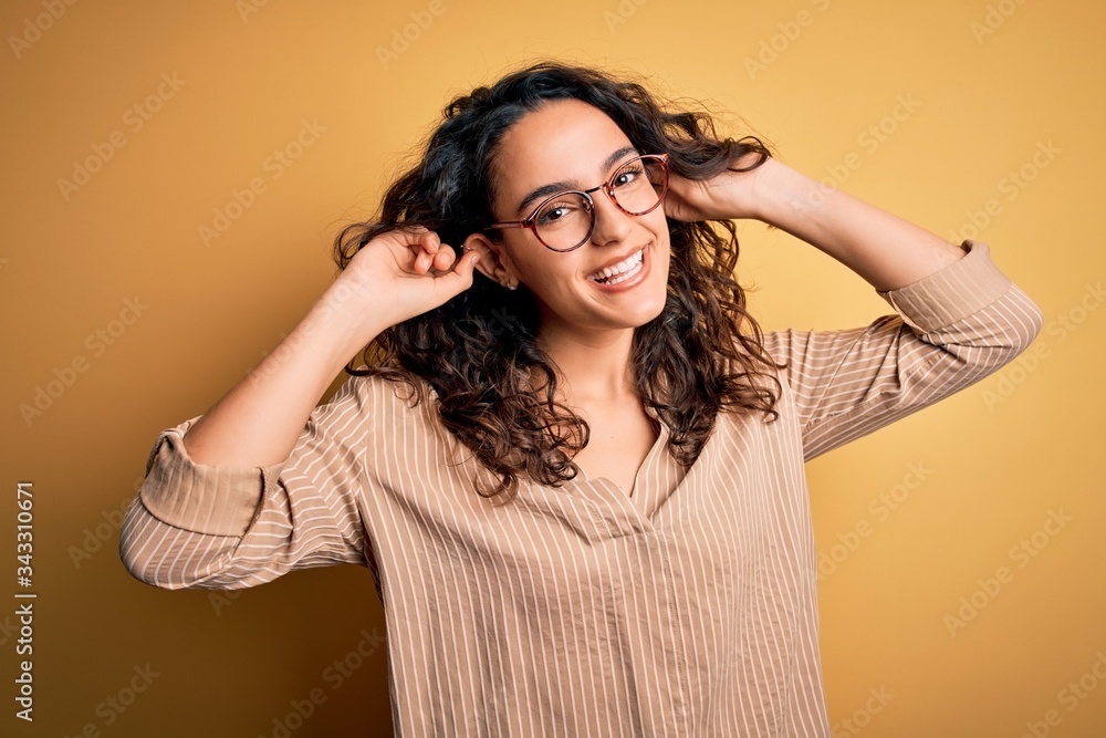 Beautiful woman with curly hair wearing striped shirt and glasses over yellow background Smiling pulling ears with fingers, funny gesture. Audition problem Beautiful woman with curly hair wearing striped shirt and glasses over yellow background Smiling pulling ears with fingers, funny gesture. Audition problem