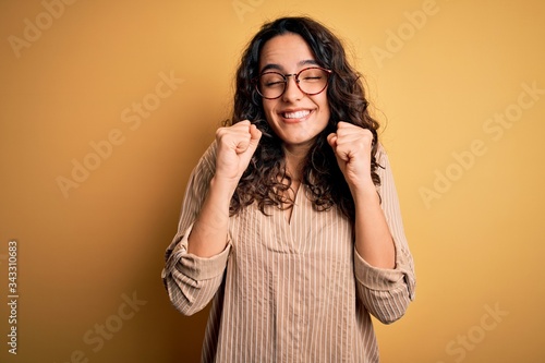 Beautiful woman with curly hair wearing striped shirt and glasses over yellow background excited for success with arms raised and eyes closed celebrating victory smiling. Winner concept.