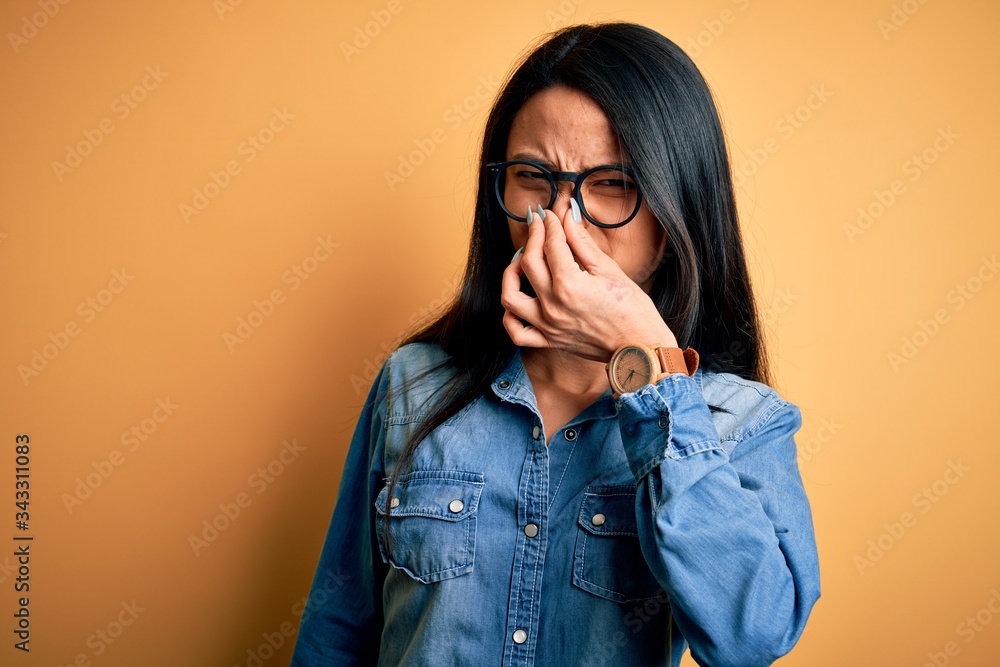 Young beautiful chinese woman wearing casual denim shirt over isolated yellow background smelling something stinky and disgusting, intolerable smell, holding breath with fingers on nose. Bad smell Young beautiful chinese woman wearing casual denim shirt over isolated yellow background smelling something stinky and disgusting, intolerable smell, holding breath with fingers on nose. Bad smell