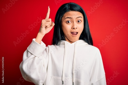 Young beautiful chinese sporty woman wearing sweatshirt over isolated red background pointing finger up with successful idea. Exited and happy. Number one.