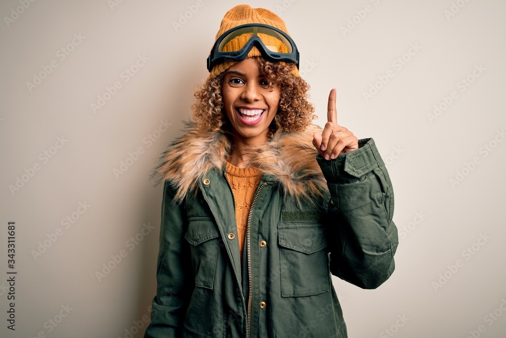Young african american skier woman with curly hair wearing snow sportswear and ski goggles showing and pointing up with finger number one while smiling confident and happy.