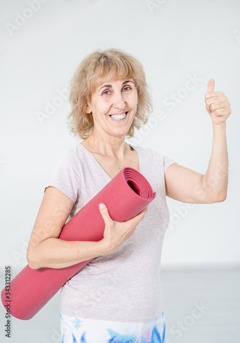 Happy senior woman holds red mat and shows thumbs up gesture on gray background
