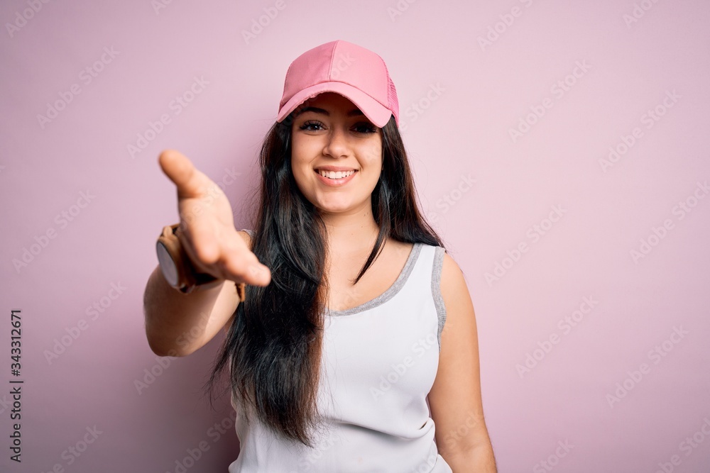 Young brunette woman wearing casual sport cap over pink background smiling friendly offering handshake as greeting and welcoming. Successful business. Young brunette woman wearing casual sport cap over pink background smiling friendly offering handshake as greeting and welcoming. Successful business.