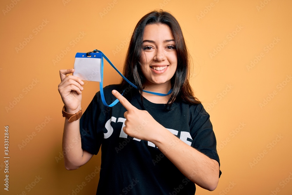 Young brunette worker woman wearing staff t-shirt as uniform showing id card very happy pointing with hand and finger