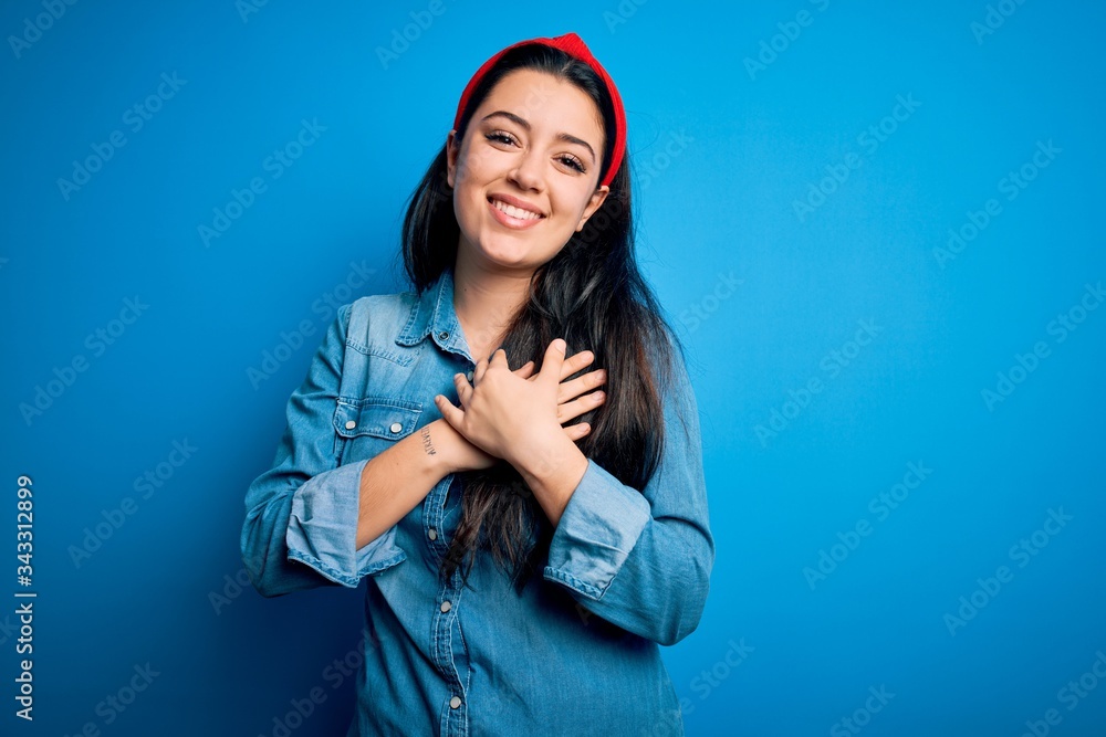 Young brunette woman wearing casual denim shirt over blue isolated background smiling with hands on chest with closed eyes and grateful gesture on face. Health concept. Young brunette woman wearing casual denim shirt over blue isolated background smiling with hands on chest with closed eyes and grateful gesture on face. Health concept.