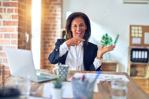 Middle age beautiful businesswoman working using laptop at the office amazed and smiling to the camera while presenting with hand and pointing with finger.