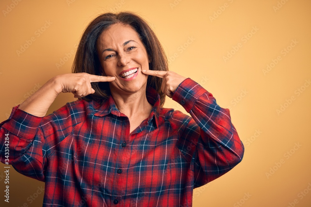 Middle age beautiful woman wearing casual shirt standing over isolated yellow background smiling cheerful showing and pointing with fingers teeth and mouth. Dental health concept. Middle age beautiful woman wearing casual shirt standing over isolated yellow background smiling cheerful showing and pointing with fingers teeth and mouth. Dental health concept.