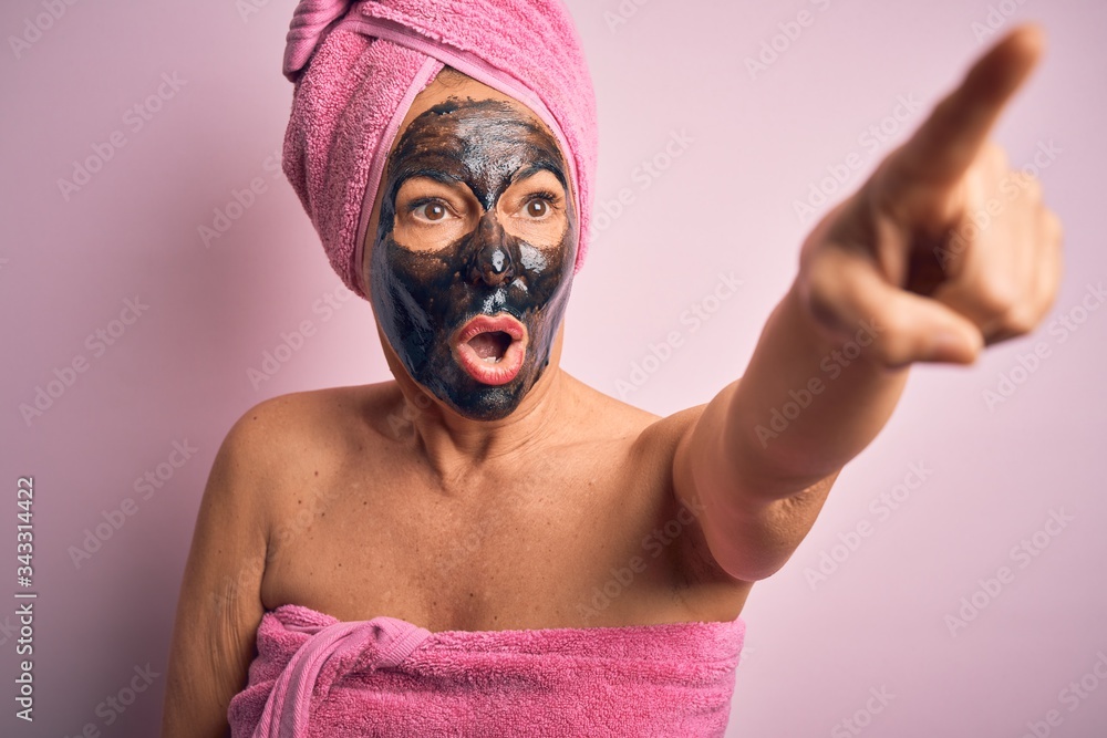 Middle age brunette woman wearing beauty black face mask over isolated pink background Pointing with finger surprised ahead, open mouth amazed expression, something on the front Middle age brunette woman wearing beauty black face mask over isolated pink background Pointing with finger surprised ahead, open mouth amazed expression, something on the front