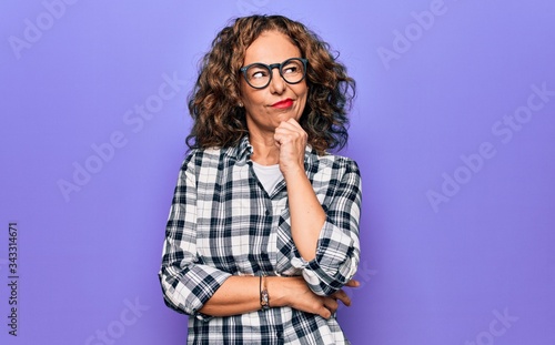 Middle age beautiful woman wearing casual shirt and glasses over isolated purple background thinking concentrated about doubt with finger on chin and looking up wondering