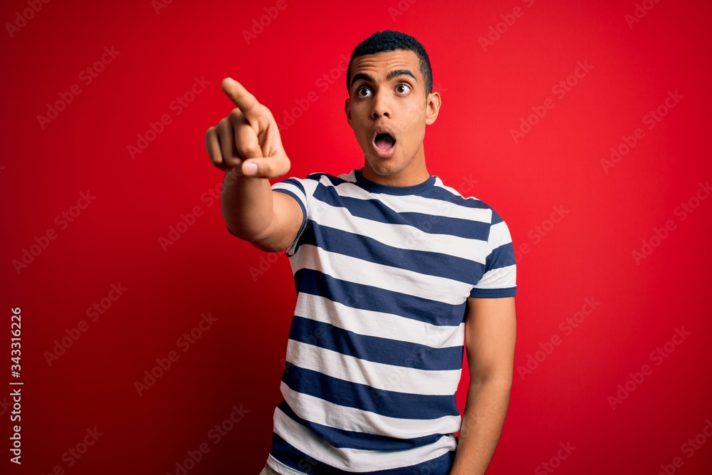 Handsome african american man wearing casual striped t-shirt standing over red background Pointing with finger surprised ahead, open mouth amazed expression, something on the front Handsome african american man wearing casual striped t-shirt standing over red background Pointing with finger surprised ahead, open mouth amazed expression, something on the front