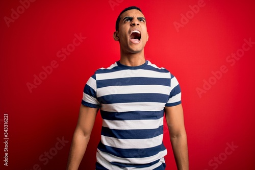Handsome african american man wearing casual striped t-shirt standing over red background angry and mad screaming frustrated and furious  shouting with anger. Rage and aggressive concept.
