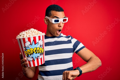 Young handsome african american man watching movie using 3d glasses eating popcorns Looking at the watch time worried, afraid of getting late
