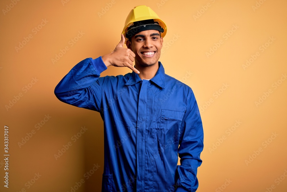 Young handsome african american worker man wearing blue uniform and security helmet smiling doing phone gesture with hand and fingers like talking on the telephone. Communicating concepts.